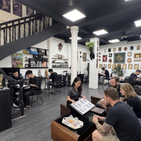 Hình ảnh chụp toàn cảnh không gian nội thất hiện đại của Qneedle Tattoo Studio với thiết kế 2 tầng, thang xoáy đen đặc trưng. Khu vực tiếp khách có sofa da nâu, nơi quản lý đang tư vấn album hình xăm cho nhóm khách nước ngoài. Phía sau là các bàn làm việc của nghệ sĩ xăm đang tập trung thiết kế và các phòng xăm riêng biệt được trang trí bằng hàng trăm bằng khen và cúp giải thưởng quốc tế. Tiệm có chi nhánh tại Hà Nội và TP. Hồ Chí Minh.