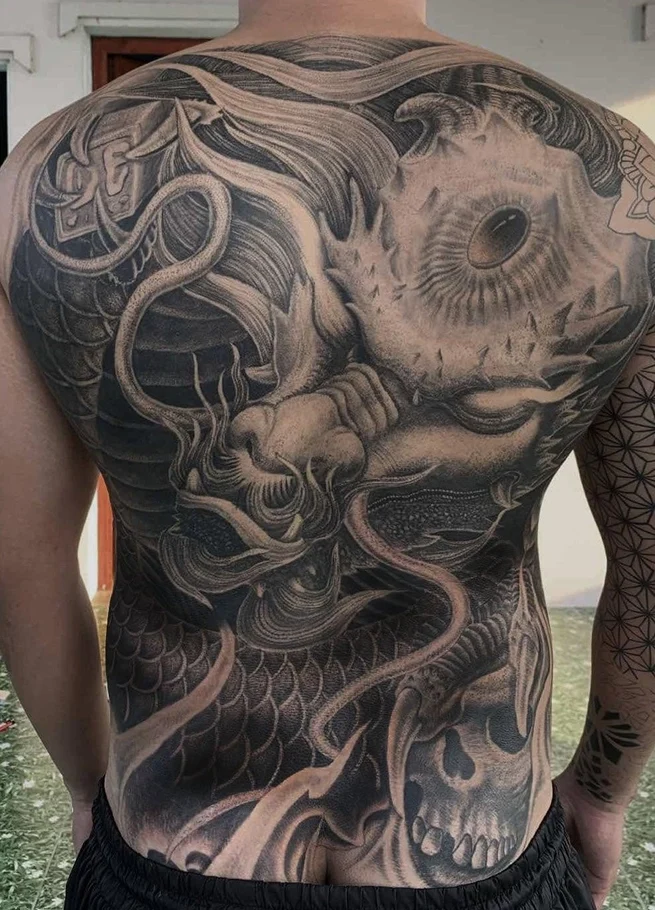 Black and grey full back tattoo featuring a stone-carved Asian dragon with intricate scales, flowing details and sculptural textures at Qneedle Tattoo Studio